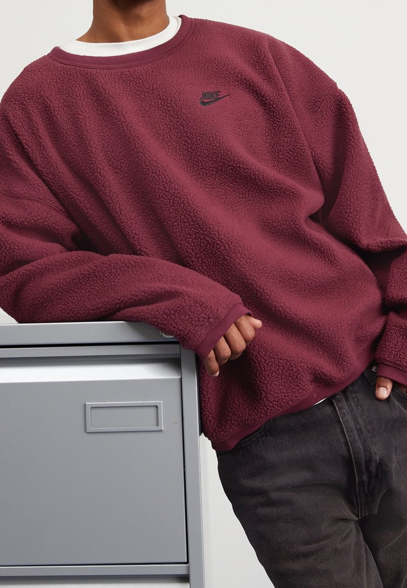 Sudadera de forro polar burdeos con superficie texturizada, cuello redondo y un pequeño logo negro de Nike en el pecho, combinada con vaqueros oscuros.