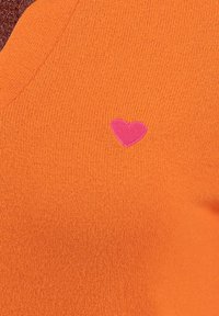 Pullover a V in morbido materiale lavorato di color arancione brillante, con un piccolo icona a forma di cuore rosa sul lato sinistro. Texture liscia con dettagli a costine.