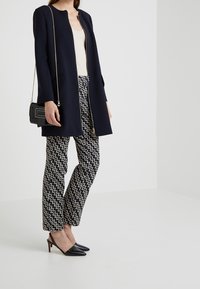 Granatowy długi płaszcz z okrągłym dekoltem, w połączeniu ze wzorzystymi spodniami w kremowe i granatowe zygzaki oraz czarnymi czółenkami na odkrytej pięcie w szpic.