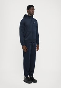 Ensemble de survêtement à capuche bleu marine avec fermeture éclair à l'avant et deux poches avant, fabriqué en tissu lisse. Associé à des baskets noires. Design simple et sportif.