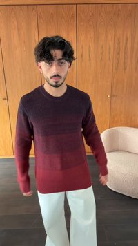 Young man with dark curly hair wearing a red to dark purple gradient sweater and white pants stands indoors with wooden cabinets and a beige armchair.