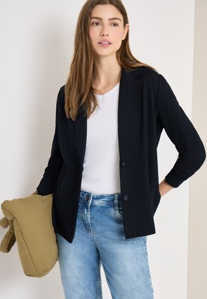 Jeune femme portant un blazer noir, une chemise blanche et un jean bleu, tenant une doudoune beige dans une main, sur un fond uni.