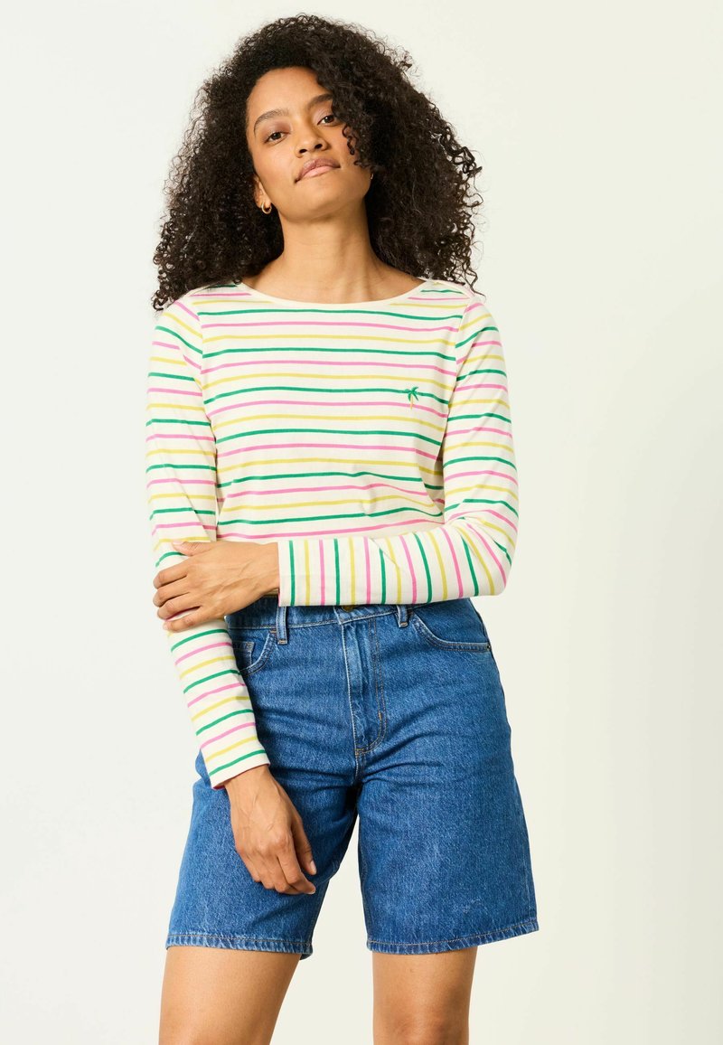 Donna con capelli ricci che indossa una maglia bianca a maniche lunghe con strisce pastello e shorts di jeans blu, in piedi davanti a uno sfondo semplice.