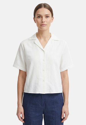 Donna che indossa una camicia bianca a maniche corte con bottoni e colletto e pantaloni blu scuro, in piedi davanti a uno sfondo uniforme.