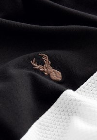 Svart tyg med en texturerad vit sektion; har ett litet broderat brunt rådjurshuvud. Mjuk, slät material med kontrasterande färger.