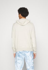 Sweat à capuche beige clair avec cordon de serrage et grande capuche. Présente des empiècements texturés aux épaules et un ourlet côtelé, assorti à un pantalon tie-dye bleu clair.