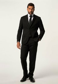 Completo nero con giacca aderente e pantaloni dritti, abbinato a una camicia bianca e una cravatta nera. Scarpe eleganti nere lucide completano il look.