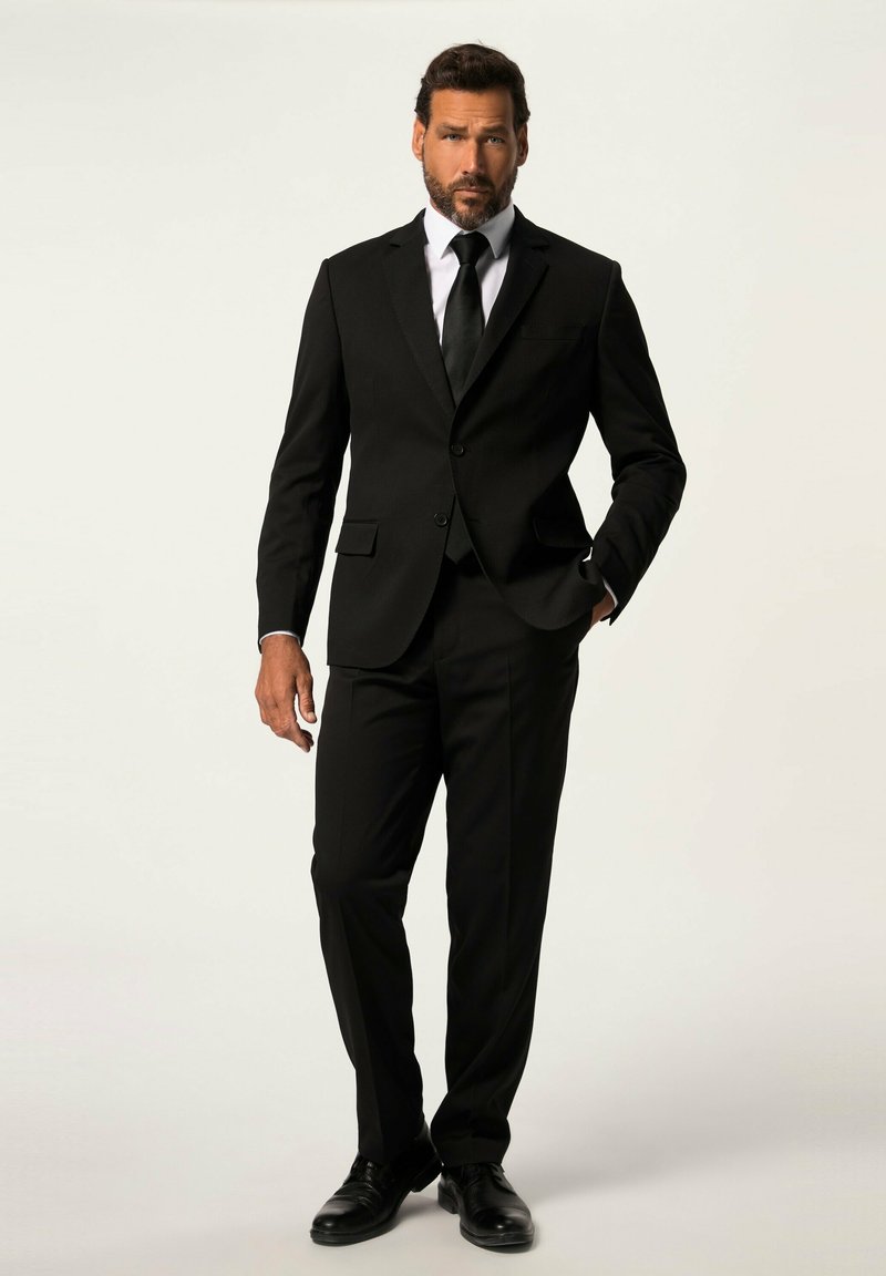 Completo nero con giacca aderente e pantaloni dritti, abbinato a una camicia bianca e una cravatta nera. Scarpe eleganti nere lucide completano il look.