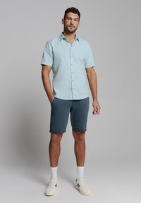 Lichtblauw korte mouwen overhemd met knopen, gecombineerd met donkergrijze shorts. Het model draagt witte sokken en crèmekleurige sneakers. Eenvoudig, casual ontwerp.