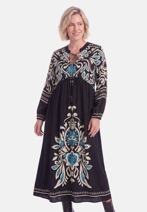 Vrouw met kort blond haar die een lange zwarte jurk draagt met grote blauw-witte bloemenpatronen en een strik bij de halslijn.