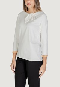 Top bianco a maniche lunghe con dettaglio a nodo sul colletto e polsini decorati in una trama contrastante chiara, abbinato a pantaloni neri.