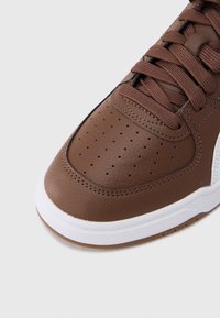 Καφέ δερμάτινο αθλητικό παπούτσι με τρύπες στο μπροστινό μέρος, λευκή λαστιχένια σόλα και καφέ κορδόνια σε αποχρώσεις που ταιριάζουν. Διαθέτει αντίθετες λευκές λεπτομέρειες στα πλάγια.