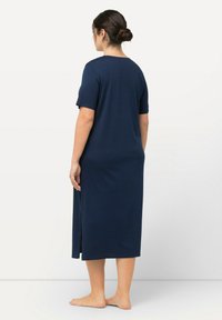 Žena s tmavými vlasy v drdolu, oblečená v námořnicky modrých šatech s krátkým rukávem a midi délkou, se bočním rozparkem, stojící bosá a sice otočená zády.