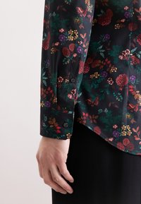Camicetta nera con motivo floreale colorato, caratterizzata da disegni rossi, verdi e viola. Maniche lunghe con polsini a bottone, texture del tessuto liscia.