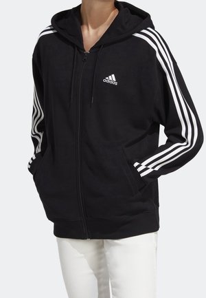 Personne portant un sweat à capuche zippé noir Adidas avec le design à trois bandes blanches sur les manches et le logo blanc Adidas, associé à un pantalon blanc.