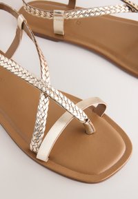 Sandali bassi marroni con un cinturino intrecciato metallico, caratterizzato da un design incrociato. La suola è liscia e di forma quadrata.