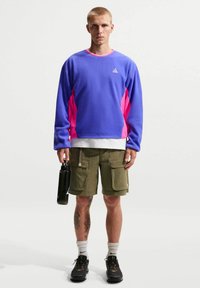 Νέος άνδρας με πουκάμισο μακρυμάνικο σε αποχρώσεις του μωβ και ροζ και πράσινα cargo σορτς κρατώντας ένα μαύρο παγούρι νερού, στέκεται μπροστά σε λευκό απλό φόντο.