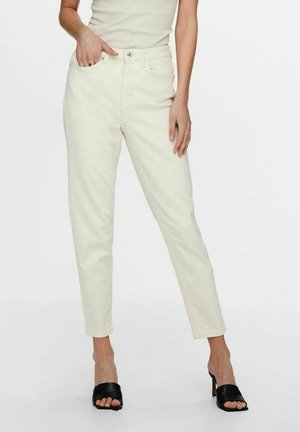 Žena nosí kalhoty s vysokým pasem, rovné nohavice v barvě slonové kosti a černé otevřené boty na podpatku, stojí s jednou rukou v kapse.