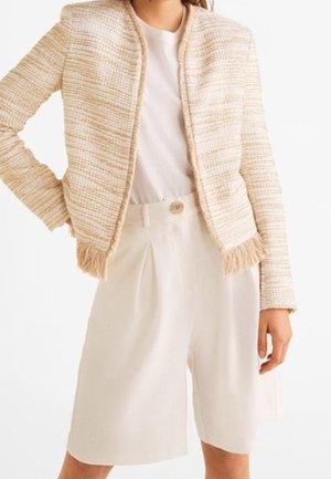 Kobieta ubrana w kremową kurtkę z tweedu z detalami z frędzlami, na białej bluzce i kremowych szortach z wysokim stanem zapinanych na guziki.