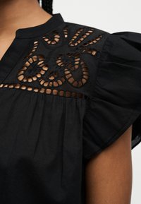 Κοντινό πλάνο σε μαύρο μπλουζάκι με περίπλοκη κεντητική με κοψίματα και κοντό μανίκι με βολάν σε βραχίονα με μέσο τόνο δέρματος.