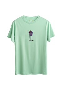 T-shirt en coton vert clair présentant un graphique central d'une personne portant un manteau violet, tenant un appareil photo, avec un pantalon et des chaussures noirs.