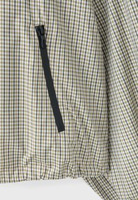 Gros plan sur un tissu de veste à carreaux verts, blancs et bleus avec une poche zippée verticale noire imperméable sur le côté.