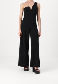 Svart jumpsuit med en axelrem, djup V-ringning, figurnära midja, vida ben och slät tyg; parat med svarta platåsandaler.