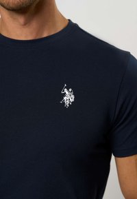 Tamsiai mėlynas medvilninis marškinėlis su mažu baltu išsiuvinėtu logotipu, vaizduojančiu polo žaidėją ant arklio. Klasikinio tipo apvalus kaklas.