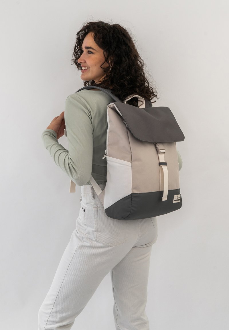 Backpack in beige and gray fabric, featuring a flap closure, adjustable straps, and contrasting black accent at the bottom.