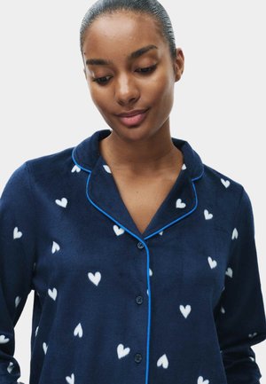 Pigiama top navy in morbido tessuto, con motivo a cuori bianchi. Rifinito con piping blu, presenta una chiusura frontale a bottoni e colletto.