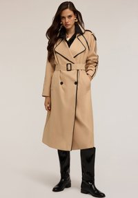 Beige trenčkot s černým lemováním, dvouřadým zap�ínáním, páskem v pase a kapsami. Černé vysoké boty doplňují celý outfit.