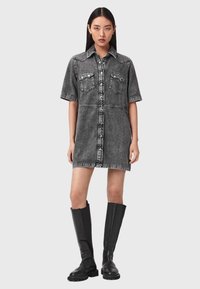 Nainen pukeutuneena kuluneen mustaan denimmekkoon, jossa on lyhyet hihat, etutaskut rinnalla ja mustat polven korkeudelle ulottuvat nahkasaappaat.