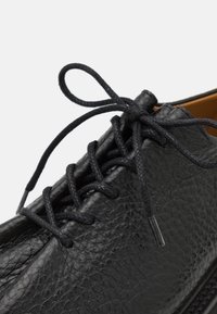 Chaussure en cuir noir avec une surface texturée, dotée de lacets ronds noirs et d'un design à profil bas. Accentuée d'un intérieur beige.