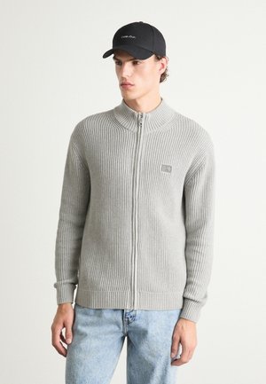 Suéter de punto gris con cremallera y diseño de canalé vertical, cuello alto y un pequeño parche con logo, combinado con unos vaqueros de mezclilla azul claro y una gorra negra.