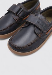 Zapatos de cuero azul marino con una correa de velcro, detalles de costura en color tostado y suela de goma. Punta redondeada y diseño casual, tipo slip-on.