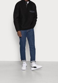 Musta fleecetakki napillisen kauluksen ja etutaskun kanssa, yhdistettynä käännettyihin sinisiin farkkuihin ja valkoisiin lenkkareihin mustilla yksityiskohdilla.