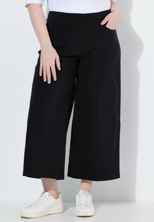 Personne portant un pantalon court noir à jambes larges et des baskets blanches, debout avec une main dans la poche sur fond blanc.