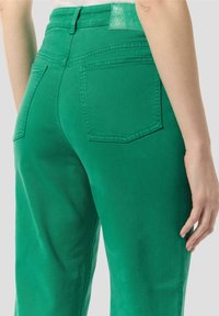 Gröna denimbyxor med hög midja, bakfickor och en texturerad grön etikett på midjebandet. Tyget har en slät finish.