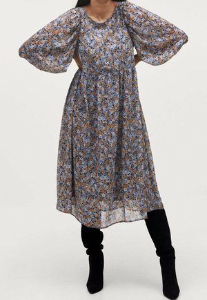 Femme portant une robe midi fleurie avec des manches longues bouffantes et des bottes noires montantes jusqu'aux genoux, debout les mains sur les hanches.