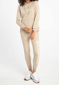 Sudadera beige con logo, joggers a juego que presentan un gran texto de la marca. La tela parece suave; zapatillas blancas con suela marrón claro.