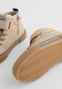 Stivaletti beige per bambini con suole in gomma testurizzata, lacci frontali e zip laterali; presenta una patch con logo e cuciture a contrasto.
