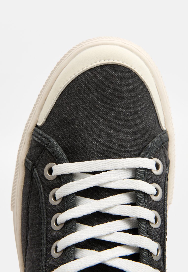 Zapatilla de lona gris con punta de goma blanca, tejido texturizado, cordones blancos y ojales de metal. Diseño minimalista y textura suave.