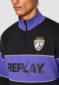 Sort zip-up sweatshirt med en lilla krave, præget af hvide striber og et stort "REPLAY" logo på brystet, plus et holdvåben med stjerner.