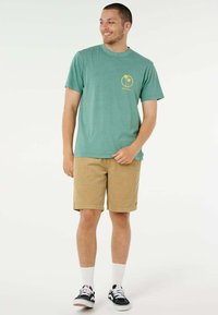 Uomo che indossa una maglietta verde con logo di palma, pantaloncini beige, calzini bianchi e scarpe da ginnastica nere, in piedi contro uno sfondo bianco.