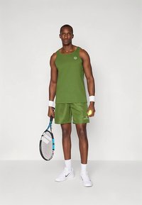 Camisola desportiva verde sem mangas e calções combinando, feitos de tecido respirável. O modelo segura uma raquete e uma bola de ténis, vestindo sapatilhas brancas.