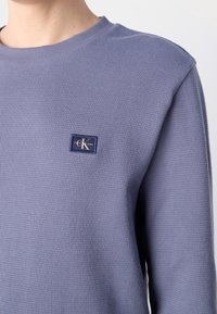 Man som bär en lila långärmad tröja i våffelstickning med en liten Calvin Klein Jeans-logotyp på vänster bröst.