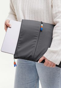 Housse d'ordinateur portable grise avec deux zippers, présentant des accents colorés sur les tirettes. Matériau en nylon lisse, tenue à côté d'un pull de couleur claire.
