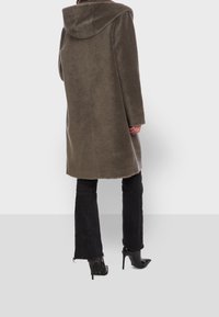 Cappotto verde oliva con cappuccio, dalla texture morbida, taglio dritto e maniche lunghe, abbinato a pantaloni neri a campana e scarpe con tacco alto.