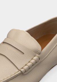 Μπέζ δερμάτινο μοκασίνι με ραμμένο λουράκι και στρογγυλεμένη μύτη, που δείχνει υφή υλικού και λεπτομερή χειροποίητη ραφή στο πλαϊνό ραφικό.