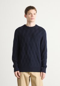 Maglione girocollo lavorato a maglia blu navy con motivo a diamante a rilievo, maniche con polsini e fondo a costine, abbinato a pantaloni beige.